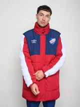 Parka larga de abrigo Umbro del Club Nacional de Football, con diseño tricolor en rojo, azul y blanco. Presenta cuello alto, cierre frontal con solapa, bolsillos con cierre en el pecho y logo de la marca junto al escudo del club.