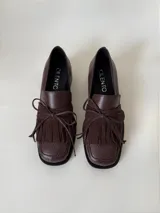 Mocasín de cuero marrón con textura granulada, diseño de horma cuadrada, detalle de flecos y lazo decorativo en el empeine, montado sobre una suela y taco bajo de color negro.