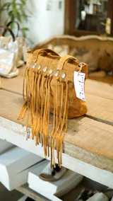 Cartera tipo bolso de cuero color tostado con diseño boho, decorada con flecos largos frontales y tachas metálicas circulares. Cuenta con doble asa de mano y cierre superior.