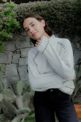 Buzo de punto color blanco con cuello alto y detalles de pespuntes decorativos en color negro en las costuras de los hombros, puños y ruedo.