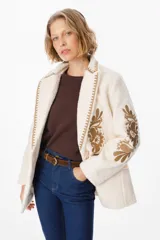 Blazer de textura suave en color crudo, con solapas clásicas y bordados florales en tonos tierra en el frente y las mangas. Presenta detalles de costura decorativa en los bordes de las solapas.