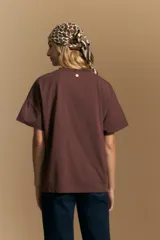 Remera de manga corta color marrón, con cuello redondo y cierre frontal parcial con botones. Presenta un diseño de calce relajado y líneas simples.