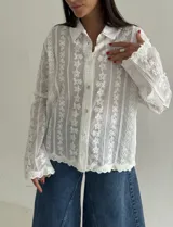 Camisa blanca de manga larga con delicados bordados florales verticales y detalles de encaje. Presenta cuello clásico, cierre frontal con botones y terminaciones festoneadas en los puños y el ruedo.