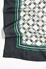 Pañuelo cuadrado de tejido suave con estampado geométrico central en tonos crema y negro, enmarcado por bordes negros con líneas decorativas en verde y celeste.
