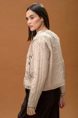 Chaqueta acolchada de color beige con bordados lineales y florales en hilo negro. Presenta un diseño de corte recto, manga larga y cierre frontal mediante lazos finos.