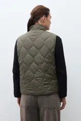 Chaleco capitoneado reversible con cuello alto, cierre frontal con botones a presión y bolsillos delanteros. Presenta un diseño bicolor en tonos verde oliva y beige con detalles en negro.