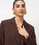 Blazer de sastrería color marrón, de corte regular y largo, con cuello peaked, cierre frontal cruzado con botones, bolsillos con tapa y mangas largas con botones decorativos.