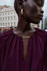 Blusa de tejido semitransparente color violeta, con cuello redondo y detalle de fornitura dorada en el escote. Presenta un diseño de escote en pico, mangas largas abullonadas con puños elásticos y corte holgado.