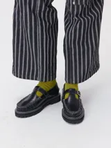 Zapato tipo guillermina de cuero negro con plataforma de goma dentada y costuras blancas a la vista. Presenta una tira con hebilla metálica sobre el empeine y diseño de puntera mocasín.