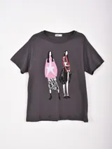 Remera gris oscuro de manga corta con cuello redondo. Presenta un estampado frontal con dos figuras femeninas estilizadas, una vestida de rosa con una estrella blanca y la otra con un look negro y rojo.
