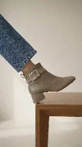 Bota corta de cuero con taco cuadrado bajo y punta redondeada. Presenta una tira decorativa en el tobillo con una hebilla metálica rectangular. Cuenta con cierre lateral interno para facilitar el calce.