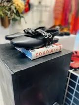 Chatas de cuero negro con punta cuadrada y talón descubierto. Presentan doble pulsera con tachas ajustables y hebilla dorada.