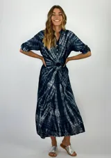 Vestido camisero midi con estampado tie-dye en tonos azul y blanco. Presenta cuello camisero, cierre frontal parcial con botones y mangas tres cuartos con trabillas. El diseño es holgado y cuenta con un ruedo asimétrico, más largo en la parte posterior.