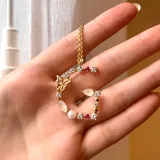 Collar dorado con dije de letra G mayúscula, adornado con perlas, piedras blancas, piedras rosas y un dije de mariposa.