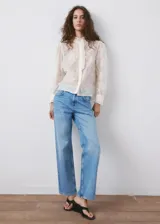 Blusa de tejido semitransparente color crudo, con diseño recto, cuello alto con volados, mangas largas abullonadas con puños abotonados y detalle de bordado floral en toda la prenda. Cuenta con cierre frontal de botones ocultos y ribete con volado en la botonera.