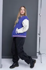 Campera universitaria violeta con mangas color crema, cierre con botones a presión, cuello, puños y ruedo acanalados con rayas blancas y violetas. Estampado frontal con la palabra "Royalty" en color verde.