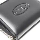 Billetera mediana de cuero vacuno color negro, con cierre perimetral y aplique ovalado con el logotipo grabado en relieve en el centro.