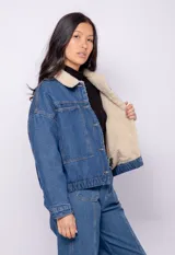 Campera de jean celeste con cuello y forro de corderito color crema, corte cuadrado, cierre frontal con botones metálicos, dos bolsillos de parche en el pecho y elástico en el ruedo.