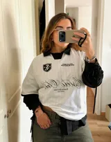 Camiseta de fútbol estilo boxy color blanco, con cuello polo y ribetes negros en las mangas. Presenta detalles estampados y bordados exclusivos en el frente y hombros.
