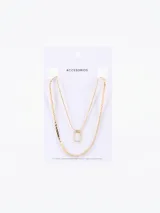 Collar doble de cadena dorada con un dije rectangular que presenta una incrustación de piedra blanca. El diseño incluye una cadena de eslabones planos y una cadena más fina, con cierre de mosquetón y cadena de alargue.