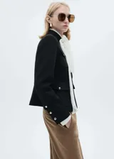 Chaqueta de estilo militar en color negro, con cuello redondo, manga larga y cierre frontal de botones metálicos dorados. Presenta cuatro bolsillos de solapa en la parte delantera y diseño de corte recto.