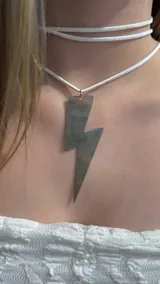 Gargantilla tipo choker compuesta por un cordón de gamuza blanca que rodea el cuello y un dije metálico plateado en forma de rayo.