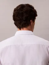 Camisa blanca de manga corta, confeccionada en tejido Oxford de algodón orgánico con elastano. Presenta cierre frontal con botones, cuello abotonado y logo de Calvin Klein Jeans bordado en el pecho.