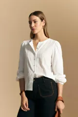 Camisa de algodón blanca con delicadas líneas bordadas verticales y textura calada. Presenta escote redondo con cierre frontal de botones, mangas largas con leve volumen en los hombros y puños elásticos.