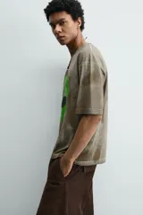 Camiseta de corte relajado con cuello redondo y manga corta. Presenta un efecto de lavado desgastado en tonos marrones y un estampado frontal de una cabeza de alienígena en color verde vibrante.