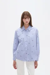 Blusa de corte holgado con cuello camisero y cierre frontal de botones. Presenta un diseño de rayas verticales finas en color azul y blanco, decorado con pequeños bordados dispersos.