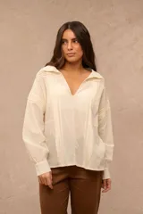 Blusa color crema de manga larga con detalles de alforzas verticales en el frente y hombros, escote en V con cuello mao y puños abotonados.