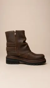 Bota de caña media confeccionada en cuero marrón con acabado desgastado. Presenta un diseño de solapa lateral con dos correas ajustables con hebillas metálicas y suela de goma dentada con plataforma.
