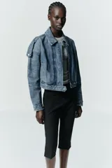 Cazadora de denim estilo crop con cuello solapa, manga larga y cierre frontal con cremallera y botón metálico. Presenta bolsillos de vivo en el delantero y detalle de trabillas en los hombros.