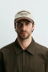 Gorra de seis paneles de algodón en color beige con visera marrón contrastante. Cuenta con un diseño bordado en marrón en el frente con texto alusivo.
