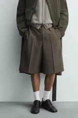 Bermuda de vestir color marrón oscuro, corte relajado y tiro alto, con pliegues frontales y cierre de botón y cremallera.