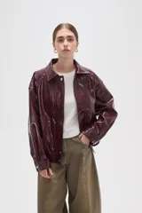 Chaqueta de eco-cuero en color bordó con acabado brillante, diseño holgado, cuello camisero, cierre frontal con botones a presión y detalle de elástico fruncido en el ruedo.