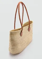 Bolso tipo cesta de playa confeccionado en hoja de palma trenzada, con doble asa de hombro larga en cuero trenzado color marrón y detalles de refuerzo en cuero en la base de las asas.