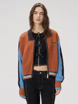 Campera estilo bomber Mutma Satifo, de diseño colorblock con cuerpo en color óxido y mangas celestes con detalles en azul marino. Cuenta con cuello camisero, cierre frontal, puños y ruedo elásticos rayados, y un parche gráfico en el pecho.