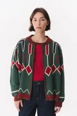 Campera tipo bomber tejida con diseño jacquard geométrico en tonos verde, rojo, azul y crema. Presenta cierre frontal, cuello redondo, puños y ruedo acanalados en color marrón, con hombros caídos y ajuste holgado.
