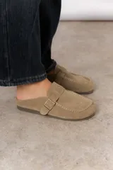 Zueco de cuero gamuzado color beige, con punta cuadrada, costuras decorativas y una tira lateral con hebilla metálica regulable. Diseño abierto en la parte trasera.