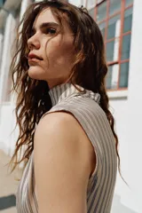 Vestido corto de cuello subido y sin mangas, con diseño fluido y pliegues en los hombros. Presenta un detalle de frunces en el frente, una abertura lateral y un borde inferior decorado con encaje negro en contraste.