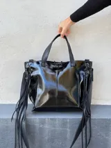 Cartera de cuero negro con acabado brillante, diseño estructurado con detalles de trenzado lateral y flecos largos. Cuenta con doble asa de mano y cierre superior.