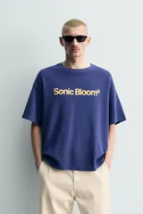 Remera de algodón con cuello redondo y manga corta, de corte holgado. Presenta un estampado frontal con la frase 'Sonic Bloom' en color amarillo.