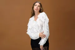 Camisa blanca de estilo romántico confeccionada en tela liviana, con volados frontales y en las mangas que aportan movimiento. Presenta escote en V y mangas largas amplias con puños ajustados.