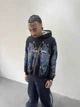 Hoodie oversize de color negro con estampado artístico en pecho y mangas en tonos azul y gris. Presenta capucha forrada con tejido a cuadros, costuras reforzadas y grifa de la marca en la manga.
