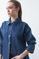 Sobrecamisa de jean negra de silueta relajada, con tachas aplicadas en líneas orgánicas que recorren la prenda. Tiene cuello camisero, cierre frontal con botones a presión y mangas largas.