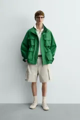 Chaqueta tipo cazadora de corte relajado en color verde, confeccionada en tejido de mezcla de algodón. Presenta cuello subido con forro interior a cuadros, manga larga con puños abotonados y cuatro bolsillos frontales de plastrón con solapa. Incluye cierre frontal de cremallera oculta por solapa con botones a presión y cintura ajustable en el interior.