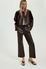 Chaqueta corta de piel color marrón, con cuello redondo, manga larga y cierre frontal con botones. Presenta un diseño con elástico en el ruedo y forro interior.