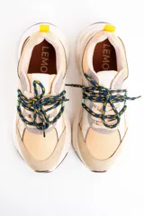 Zapatilla urbana de diseño chunky, con paneles combinados en tonos beige, arena y gris. Presenta cordones jaspeados en azul y amarillo, tirador trasero amarillo y suela de plataforma dentada en color crema.