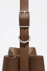 Bolso de hombro tipo cubo de estructura rígida, color marrón oscuro, confeccionado en piel vacuna. Posee asa regulable con hebilla metálica y cierre de cordón en la parte superior. Lleva el logo metálico de Bimba y Lola en la parte frontal inferior.